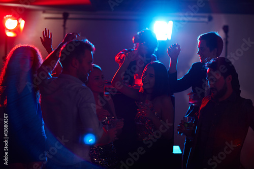 Young stylish girls and guys enjoying disco party, dancing and drinking while famous DJ mixing music on background