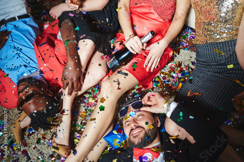 Tipsy multiethnic group of friends relaxing on floor with colorful confetti while celebrating holiday together