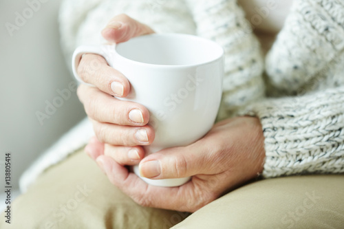 Elderly woman in warm home clothing sitting on couch and holding white cup of coffee in hands, close-up shot