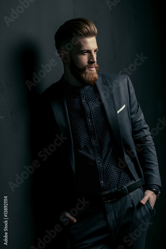 Stylish handsome bearded man in trendy suit standing with hands in pockets on black