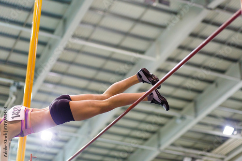 Young athletic woman vaoulting over bar with pole against flag on indoor track and field championship