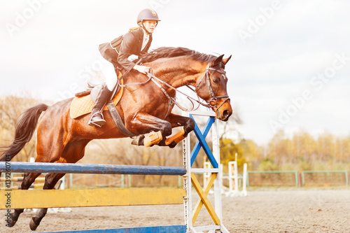 Bay horse with rider girl jump over hurdle on show jumping competition