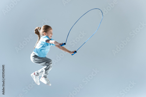 side view of happy girl humping exercising with skipping rope on grey