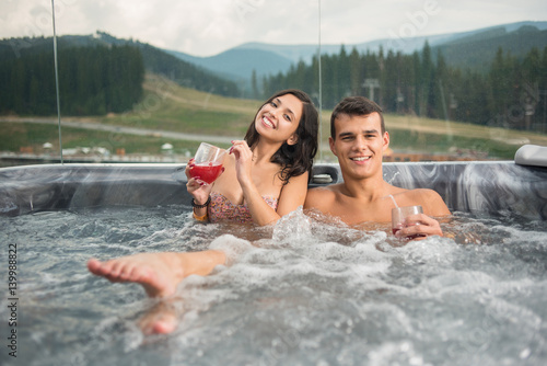 Happy young couple enjoying a bath in Jacuzzi while drinking cocktail outdoors on romantic vacation against blurred background of nature