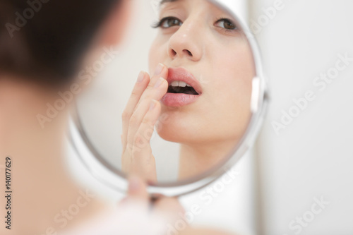 Young woman with cold sore looking in mirror at home