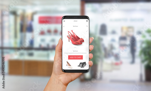 Woman buy red shoes online with mobile phone. E shopping with web site or app. Clothes and footwear shop in background.