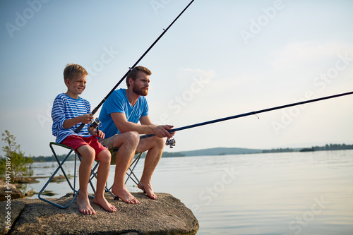 Portrait of handsome bearded father fishing with son sitting on rock in still lake with fishing rods and gear on sunny summer day