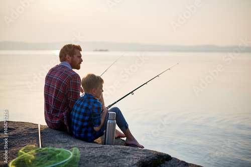 Widok z tyłu portret dorosłego mężczyzny i nastoletniego chłopca siedzącego razem na skałach łowiących pręty w spokojnych wodach błękitnego jeziora o zmierzchu, obaj ubrani w kraciaste koszule