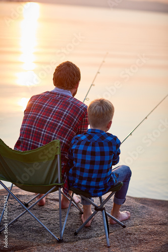 Back view portrait of father and son sitting together on rocks fishing with rods in calm lake waters in sunset light, both wearing checkered shirts