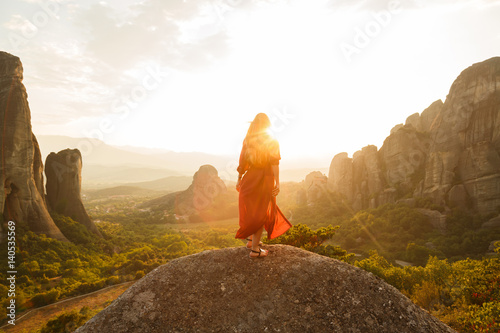 Girl in red flying dress looking at majestic sunset in Meteora valley, Greece