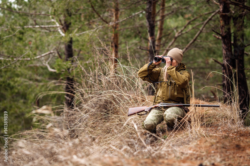 Female hunter in camouflage clothes ready to hunt, holding gun and walking in forest.