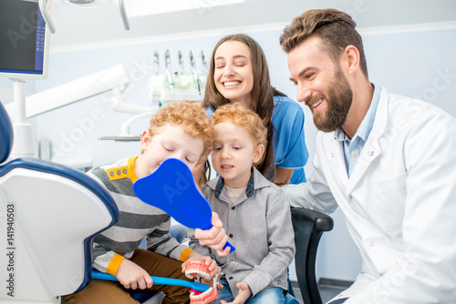 Young boys looking at the mirror with toothy smile sitting with dentist and woman assistant at the dental office