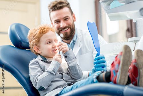 Young excited boy looking at the dental mirror sitting on the chair with dentist at the dental office