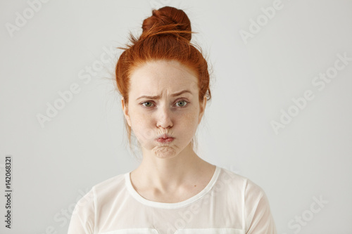 Headshot of attractive funny young female with ginger hair dressed in white blouse feeling displeased or uncomfortable with something, blowing cheeks and frowning. Human face expressions and emotions