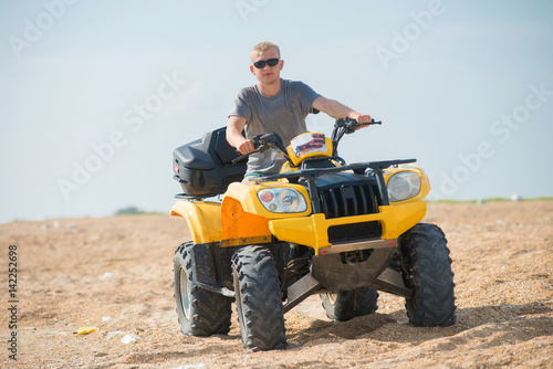 A young guy in sunglasses is riding an yellow ATV on the beach