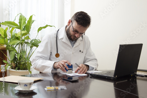 Lekarz w białym kitlu siedzi przy stole i przystawia pieczątkę do dokumentu. Ręce lekarza przystawiającego pieczątkę na recepcie. Lekarz wypisuje receptę przy biurku.