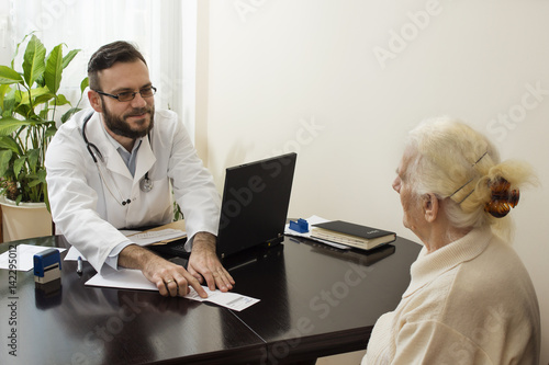 Lekarz podczas pracy. Pokazuje palcem i tłumaczy pacjentce zapis na recepcie. Stara kobieta u lekarza geriatry.