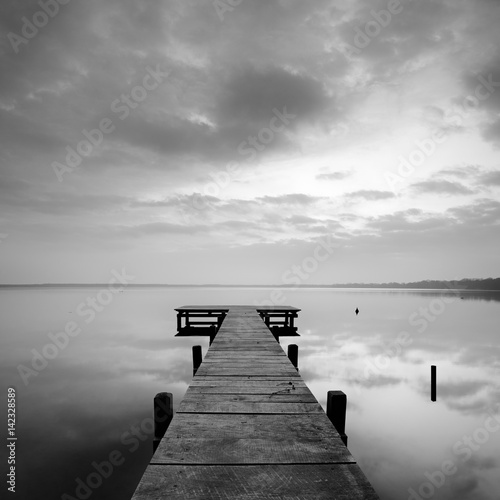 Stiller See mit Steg bei Sonnenaufgang, wolkiger Himmel, schwarz-weiß