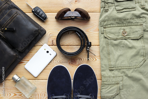 Young man accessories for spring or summer season in casual style, jeans, shoes, belt, eyeglasses, bag, mobile phone, car key and perfume on brushed wood background, fashion industry 
