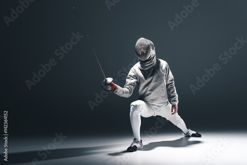 Professional fencer in fencing mask with rapier standing in position on grey