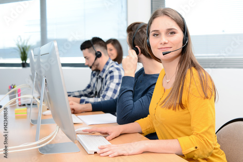 portrait of beautiful and cheerful young woman telephone operator with headset working on desktop computer in row in customer service call support helpline business center