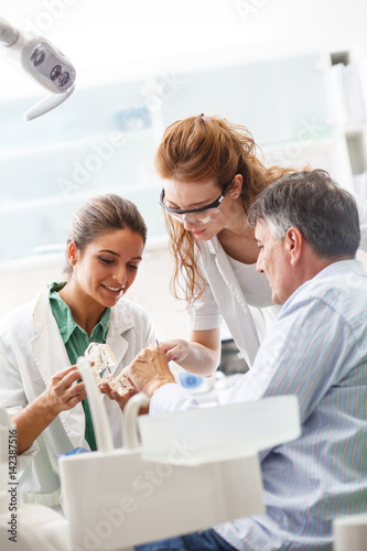 Dentist and her female assistant in dental office talking with senior patient and preparing for treatment.
