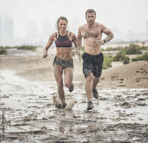 Muscular male and female athlete covered in mud running down a rough terrain with a desert background in an extreme sport race 
