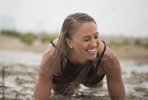 Strong athletic woman laughing with mud on her face crawling in mud or extreme sport with clean background
