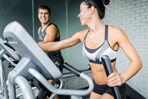 Portrait of beautiful sportive brunette woman exercising using elliptical machine next to fit man and smiling to him during workout in modern gym