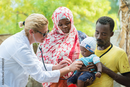 Female Caucasian doctor listening breath and heart beat of little African baby with stethoscope.Father holding the baby, mother looking at baby