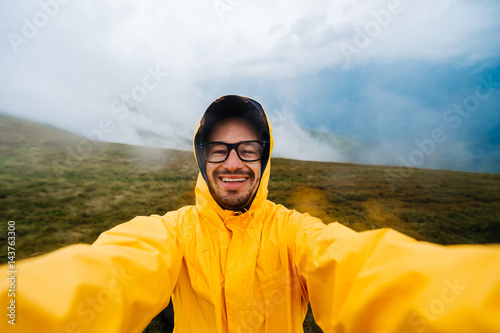 Selfie portrait of a smiling and laughing traveller man in yellow raincoat and glasses in the clouds mountains in stromy weather with rain