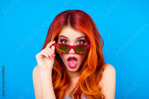 Surprised shocked girl with beautiful red curly long hair holding sunglasses and open mouth while standing on blue background