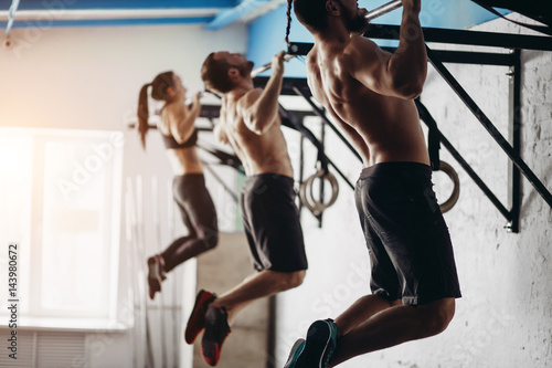 Group of tree attractive young male and female adults doing pull ups on bar in cross fit training gym with brick walls and black mats