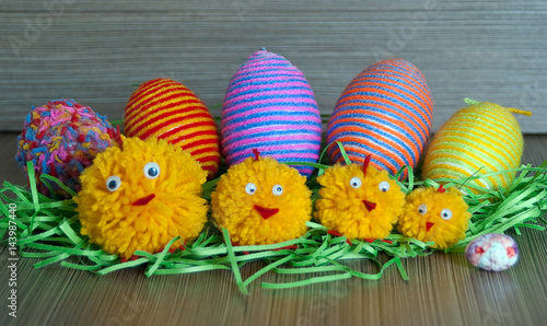 Easter decorative multicolored eggs with wrapped yarn and cute little pompom chickens in green grass nest on wooden background, close up. Easter concept. Homemade decoration. 