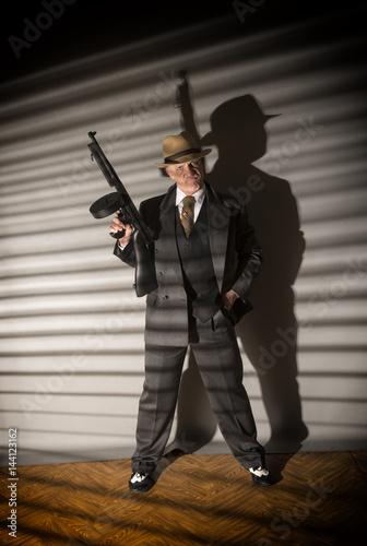 Mature man dressed as a 1940s gangster holding a machine gun, on a grey background 