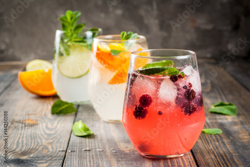 Selection of three kinds of gin tonic: with blackberries, with orange, with lime and mint leaves. In glasses on a rustic wooden background. Copy space 