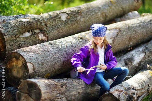 Cute little girl sitting on tree logs using a pocket knife to whittle a hiking stick