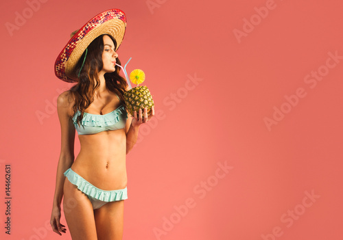 young beautiful woman in bikini and sombrero drinking exotic tropic cocktail isolated on pink background