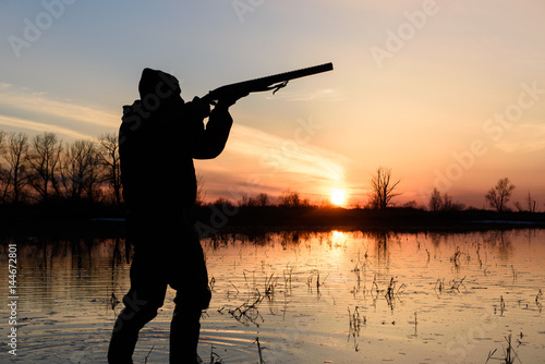 Silhouette of a hunter at sunset in the water with a gun. 