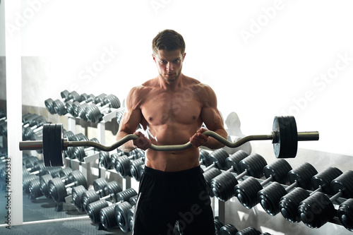 Horizontal shot of a shirtless muscular athletic young man doing biceps curls exercising with EZ curl bar at the gym workout fitness toning bodybuilding muscles sport activity concept.