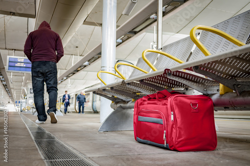 Terrorism and public safety concept with an unattended bag left under chair on platform at train station or airport and man wearing a hoodie walking away from the suspicious item (possibly terrorist)