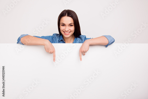 Cheerful cute girl is standing behind the white blank banner and pointing down at a copyspace