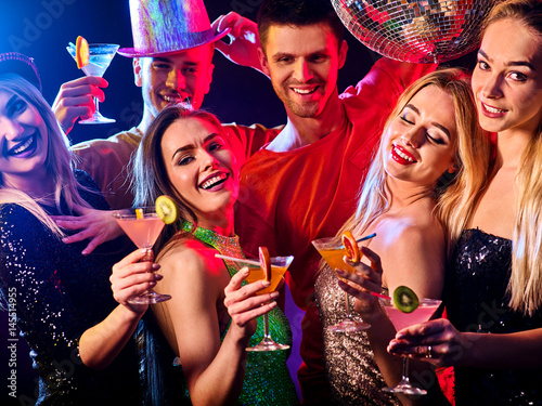 Dance party with group people dancing . Women and men have fun and drinking martini cocktail with citrus fruit in night club. Happy girl on foreground and disco ball on background. Guys wearing hats.