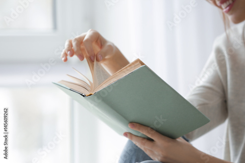 Beautiful young woman reading book near window at home, closeup