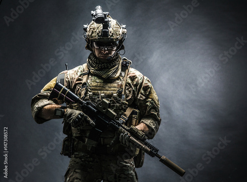 Army soldier in Combat Uniforms with assault rifle, plate carrier and combat helmet are on, Shemagh Kufiya scarf on his neck. Studio shot, dark background