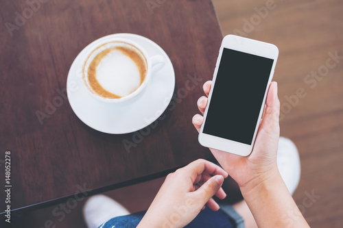 Mockup image of hands holding white mobile phone with blank black screen with coffee cup on wooden table and floor background