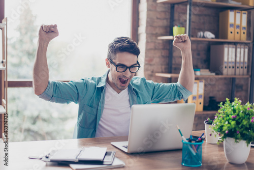 Excited screaming young man looking at the screen of his computer and triumphing with raised hands