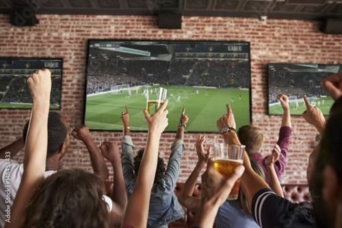 Rear View Of Friends Watching Game In Sports Bar On Screens