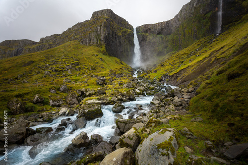 Cold water in Iceland. Waterfall in rocky mountains. Fresh and green grass. Beautiful mountain range in the backgrond.