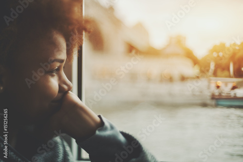 True tilt shift view of smiling black teenager girl near window traveling by ship, biracial young female is laughing and covering her mouth with hand while sitting inside of small cafe near water
